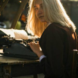 blonde haired woman in black jacket turning to look at the camera as she types a letter on a typewriter