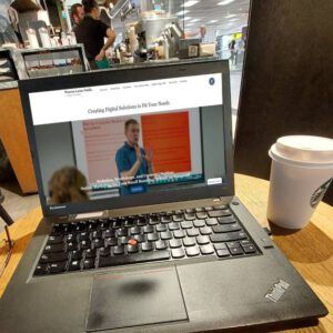 laptop on a table in a starbucks with a coffee cup to the right. on the screen of the laptop a website.